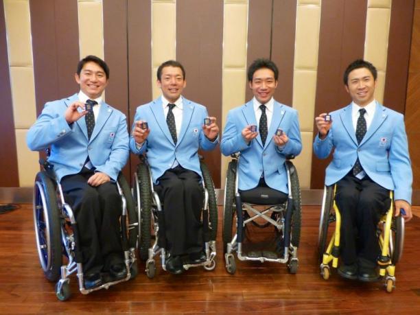 Four men in wheelchairs all wearing blue jackets show pins to the camera, smiling
