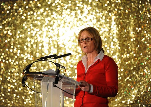 Penny Briscoe woman in red standing on stage in front of a gold background and addressing the crowd
