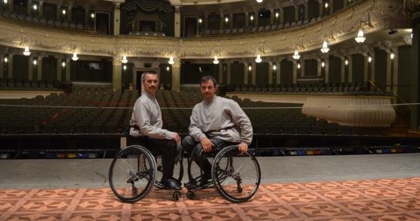 Two men in wheelchairs on a stage in front of empty seats