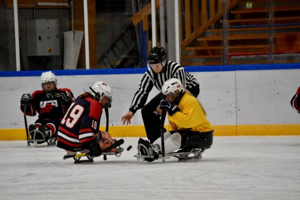 Two female sledge hockey players on the ice, fighting for the puk from the referee