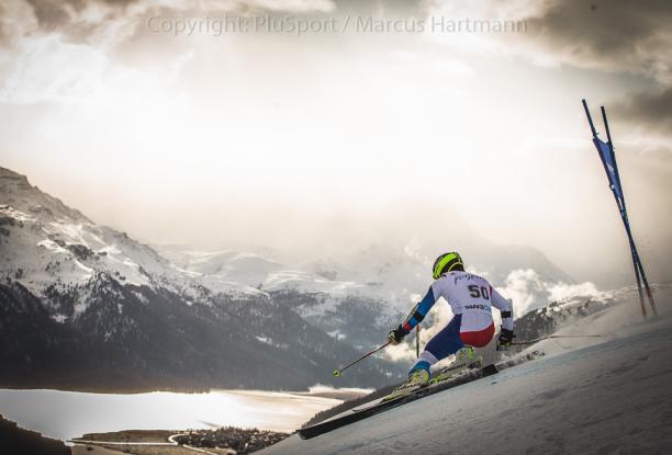 A standing alpine skiier goes down a slope with mountainous backdrop 