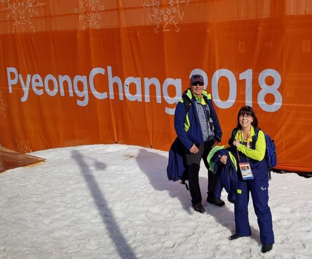 Two winter sport officials pose in front of a PyeongChang 2018 banner
