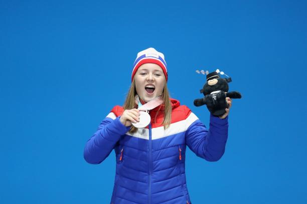 female Para Nordic skier Vilde Nilsen standing on the podium smiling holding up a silver medal and a black bear mascot