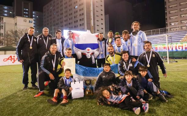 A group of male blind footballers stand with a blindfolded mascot holding a trophy and the Argentinian flag