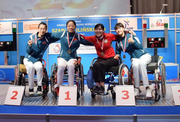Four female wheelchair fencers put arms around each other and pose on a podium