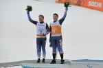 Two men in skiing suits waving flower bouquets. They both stand on a podium in the snow