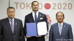 Three men standing next to each other, looking at the camera. Background: Tokyo 2020 logo. Athletic man in the middle showing a document to the camera