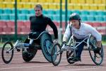 Two male athletes in racing wheelchairs on track, smile to the camera