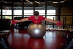 An athlete lies on his stomach on a stability ball with both his arms stretched out to the sides holding weights.
