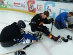 Two men in Para ice hockey sledges, one helping another get in the sledge