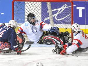 A picture of man in a sledge shooting a puck between two players to score a goal in ice hockey match