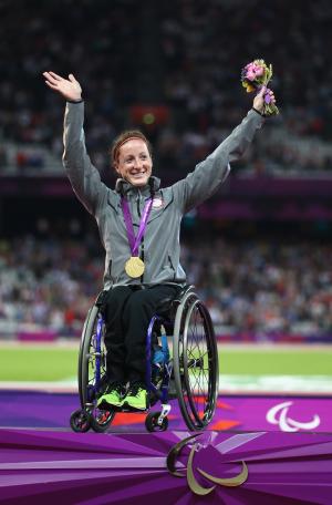 A picture of a woman on a podium with the medal around her neck and with her hands up